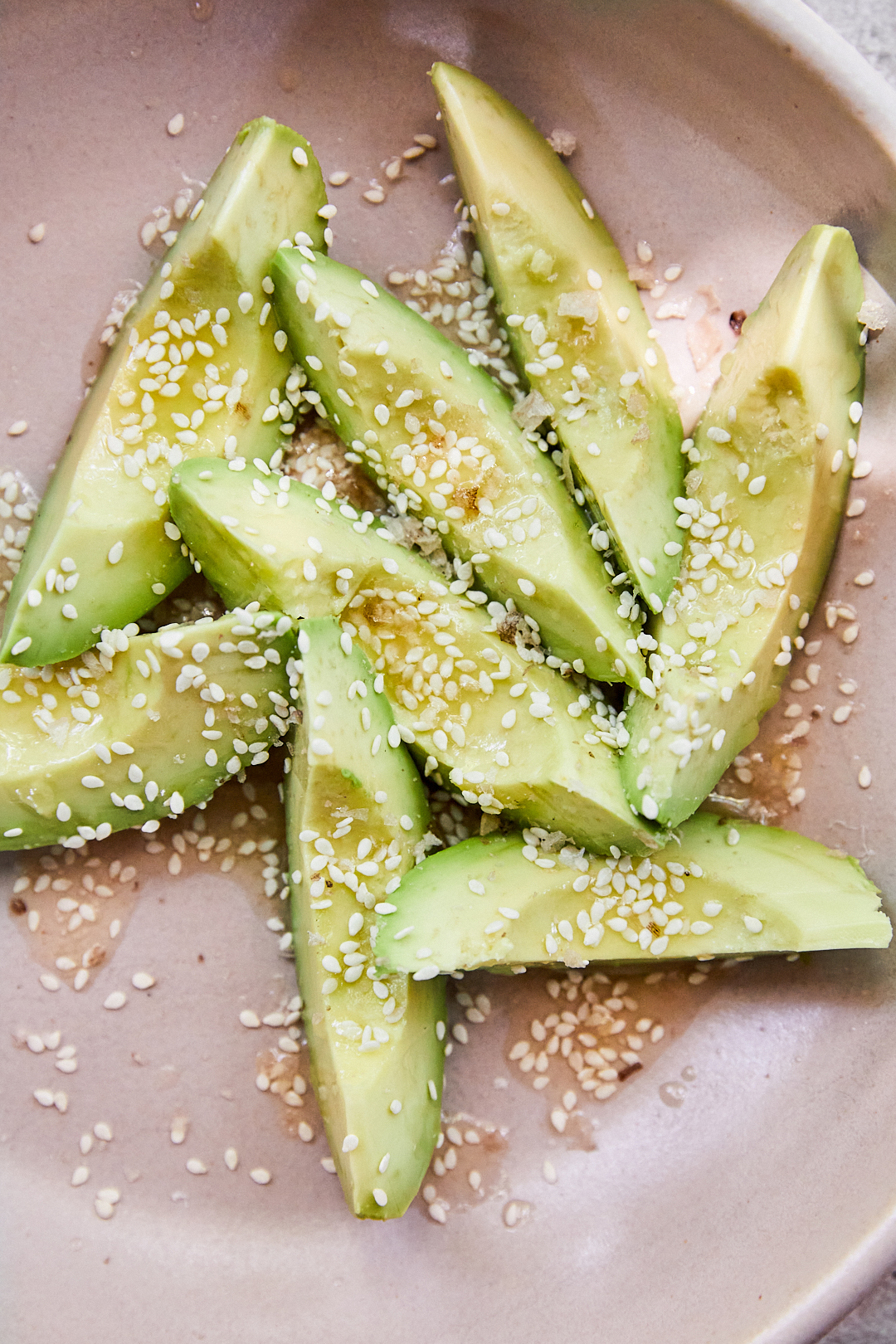A blush bowl containing wedges of avocado that are topped with sesame seeds, vinegar butter, and pieces of finishing salt. 