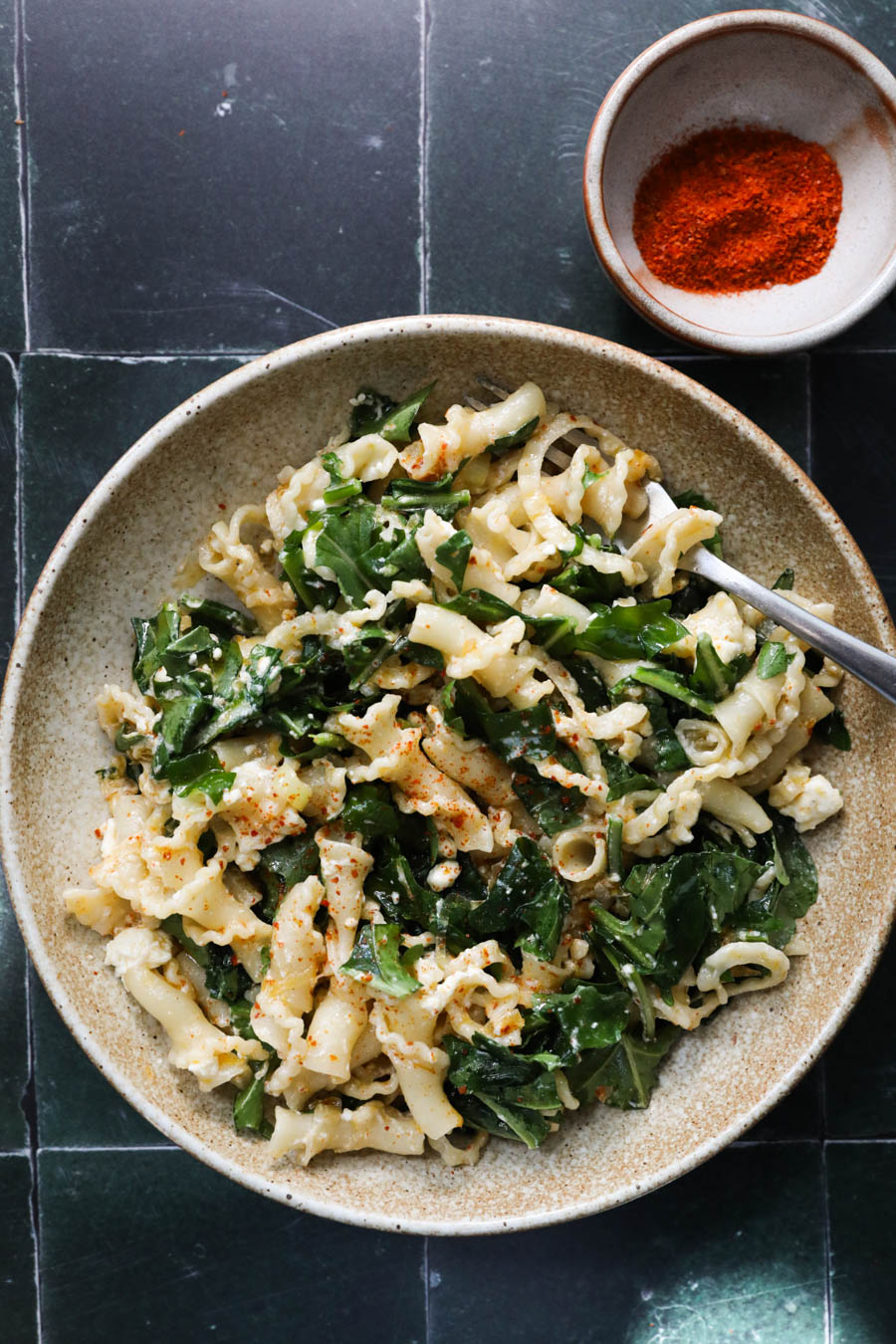 Image of pasta salad with arugula and bright red chile powder on a green tile background.