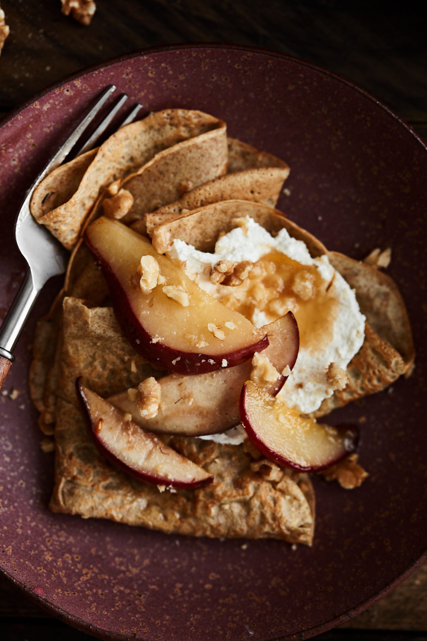 Close-up of rye crepes on a maroon plate topped with pears, whipped crepe, and walnuts. 