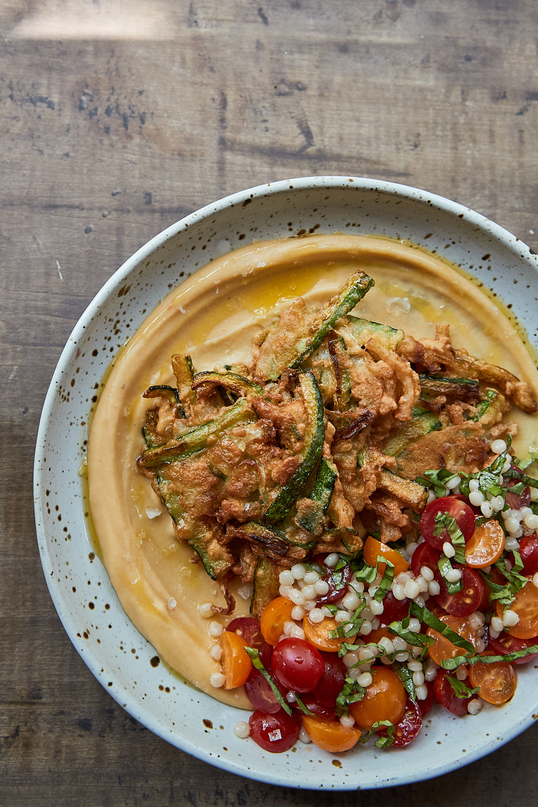 A white speckled bowl with a swoosh of creamy lentil puree topped with crispy zucchini fritters and fresh cherry tomatoes. 